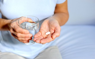 Eine sitzende Frau hält ein Wasserglas und eine Tablette in der Hand.