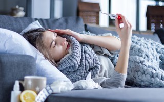 Eine Frau liegt auf der Couch und hält ein Fieberthermometer in der Hand.